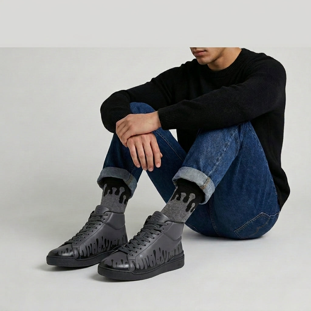 A person sitting cross-legged on a light gray studio floor, wearing 33MB Nighty mid-top dark grey statement sneakers with the signature Zero Gravity® motif of upward drops representing hope and 33MB CoZyG Eclipse gray socks with Zero Gravity® design, blue jeans, and a black long-sleeve top.