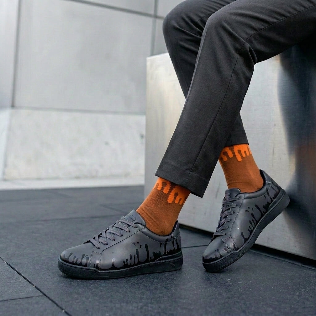 A model wearing 33MB Solar socks highlighting the symbolic orange drops against a classic camel base, styled with grey pants and nighty low 33MB premium leather sneakers featuring the Zero Gravity® signature motif symbolizing hope.