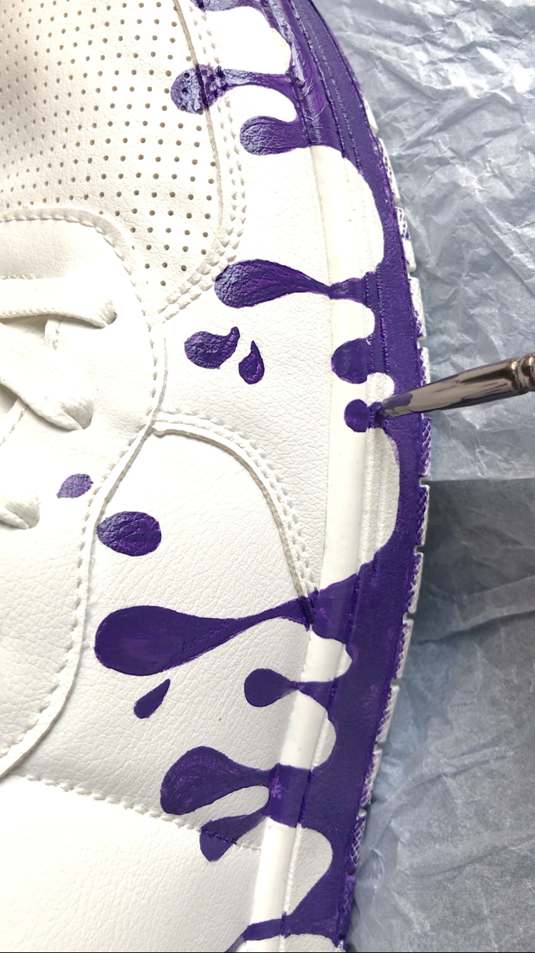 White sneaker with purple paint upward-drops being painted on a gray surface.