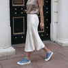 Person wearing a taupe top and white skirt with 33MB low-top Zephyr denim blue recycled canvas sneakers featuring the Zero Gravity® upward-drop motif representing hope  with white soles,  in front of a black door.