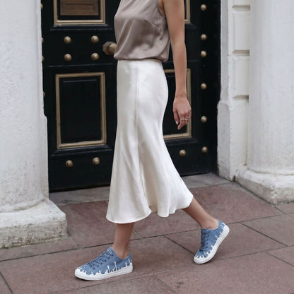Person wearing a taupe top and white skirt with 33MB low-top Zephyr denim blue recycled canvas sneakers featuring the Zero Gravity® upward-drop motif representing hope  with white soles,  in front of a black door.