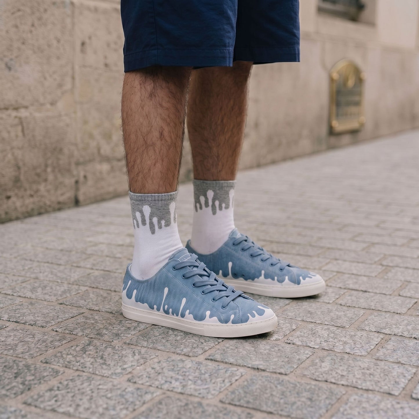 A person wearing dark shorts, 33MB low-top Zephyr denim blue recycled canvas sneakers and 33MB Lunar organic cotton CoZyG socks featuring the Zero Gravity® upward-drop motif representing hope,  standing on a stone pavement.
