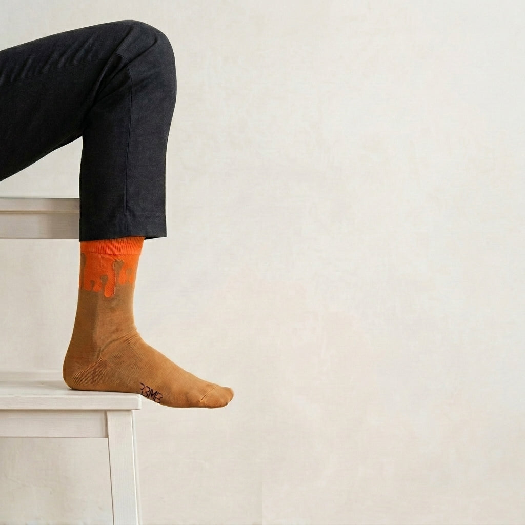 A model wearing 33MB Solar socks, highlighting the symbolic ero Gravity® signature motif of ascending drops moving upward symbolizing hope,  orange against a classic camel base and styled with dark pants
