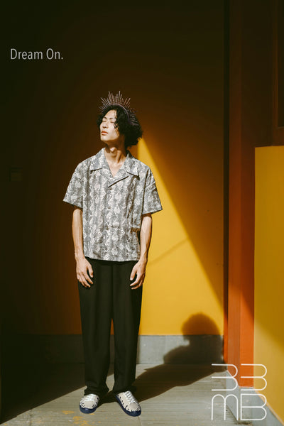 Person wearing a patterned shirt, dark pants and beige leather sneakers with black drop design standing against a warm-toned wall with 'Dream On.' text.