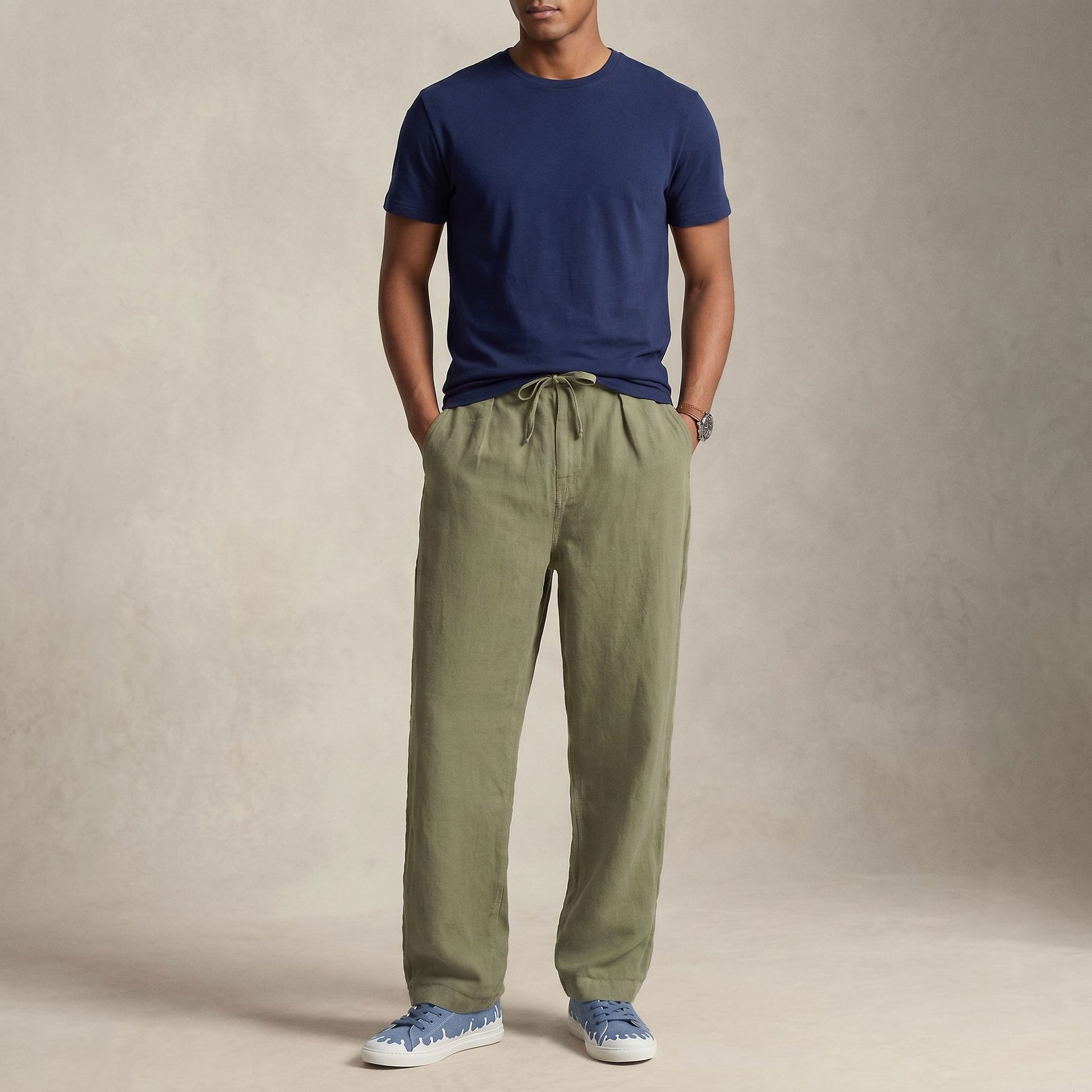A man wearing olive green drawstring pants, a navy blue t-shirt, and 33MB low-top Zephyr blue denim recycled canvas sneakers featuring the Zero Gravity® upward-drop motif representing hope  with white soles,  standing against a textured beige studio backdrop.
