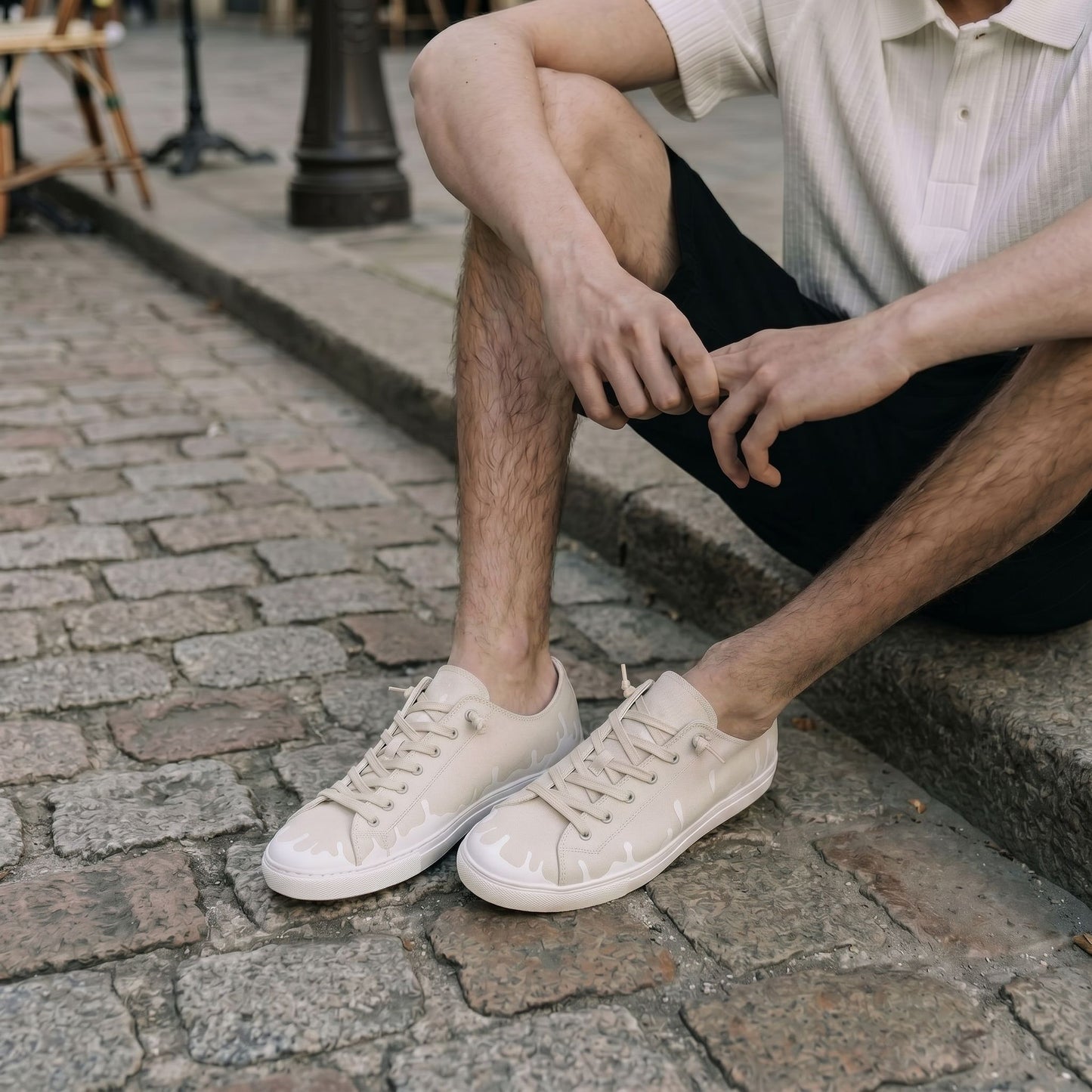 A person sitting on a stone curb wearing black shorts, a white short-sleeve ribbed polo, and off-white cream low-top 33MB sneakers featuring the Zero Gravity unique upward drip motif design near the soles representing hope. 